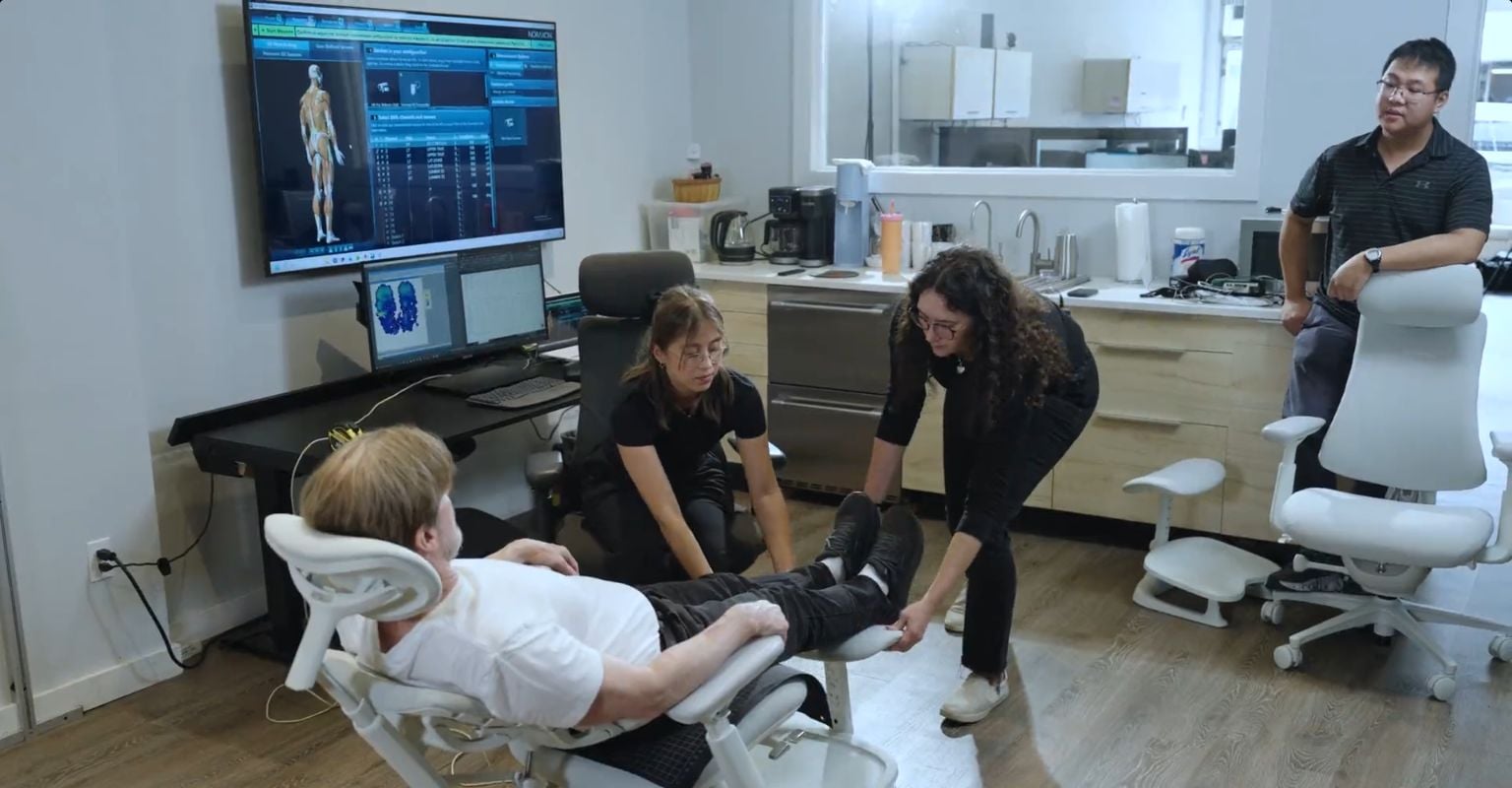 Researchers working around a participant reclined in a light-colored LiberNovo Omni Pro, with measurement screens and a second chair visible in the laboratory.